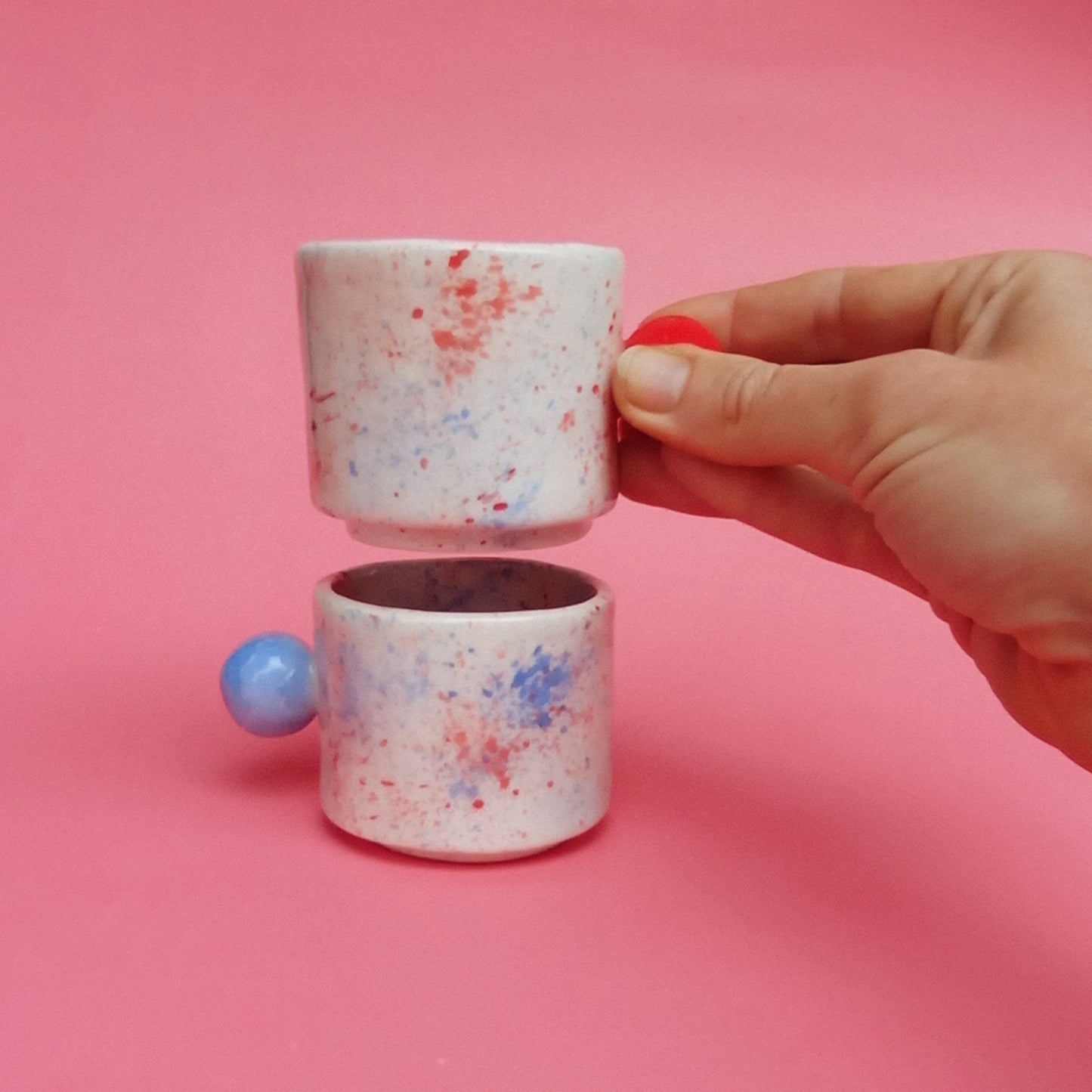 Hand holding a small ceramic cup with colorful splatter design against a pink background
