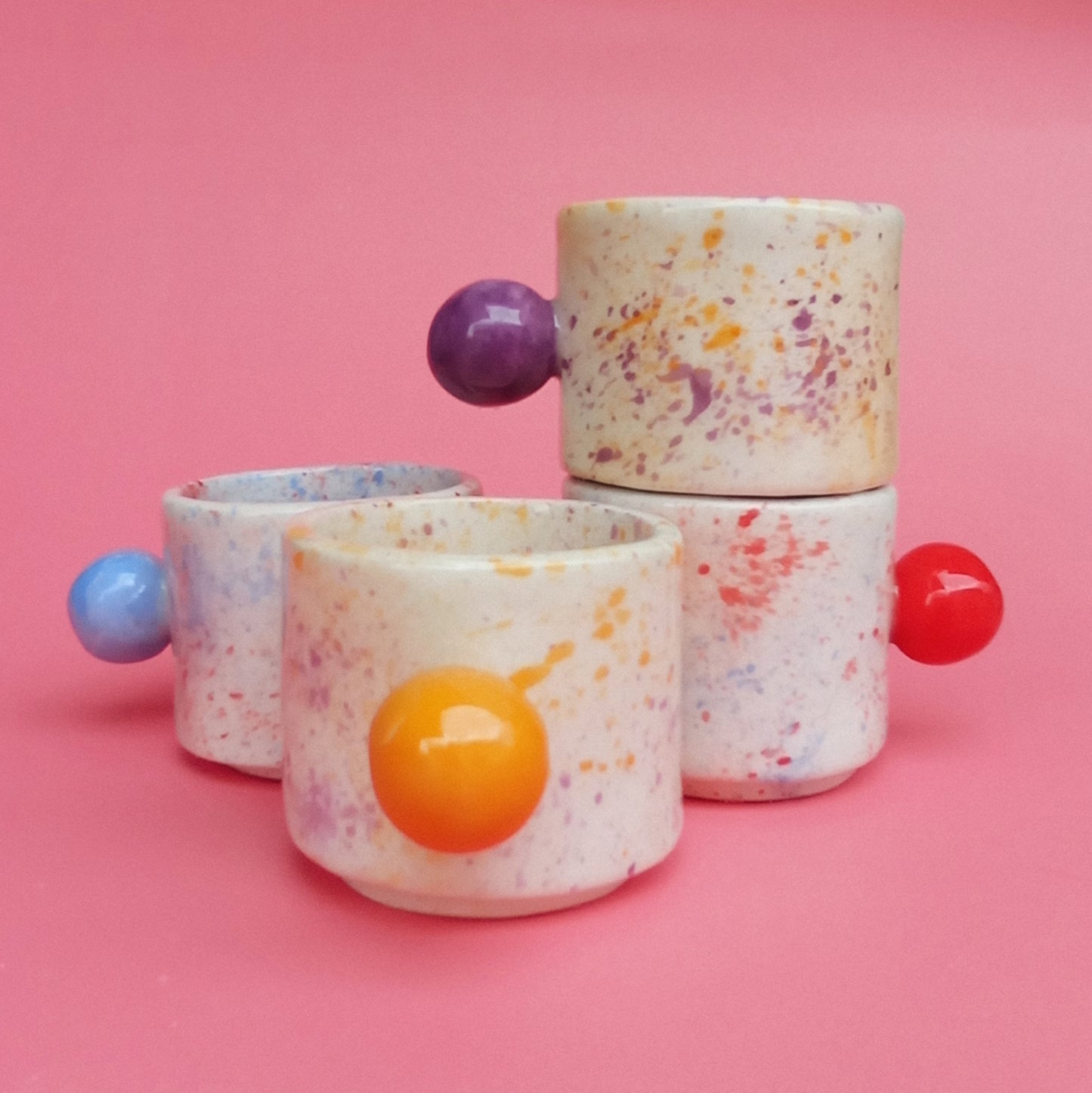 Speckled ceramic cups with colorful spherical handles on a pink background