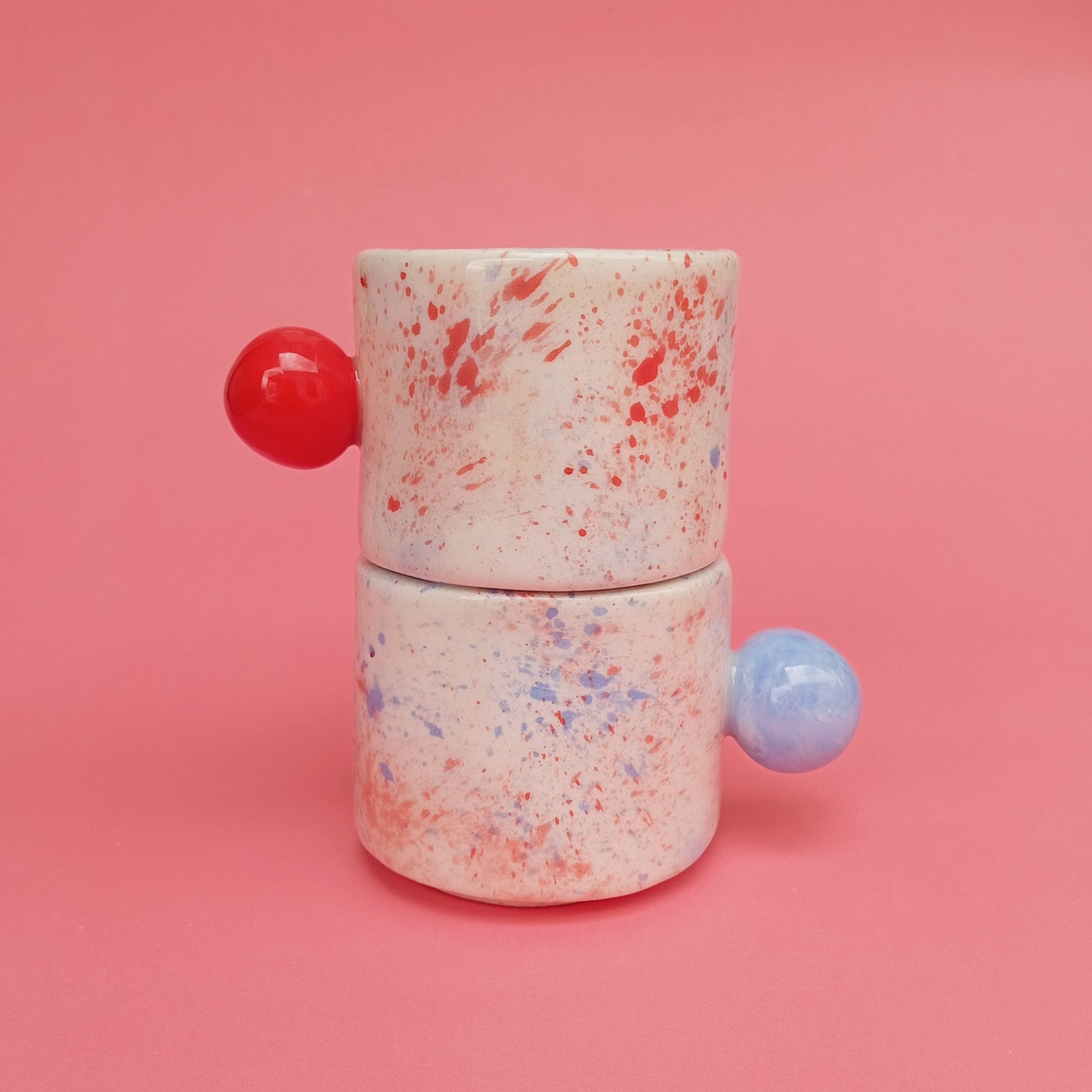 Two ceramic containers with colorful splatter design and round handles on a pink background