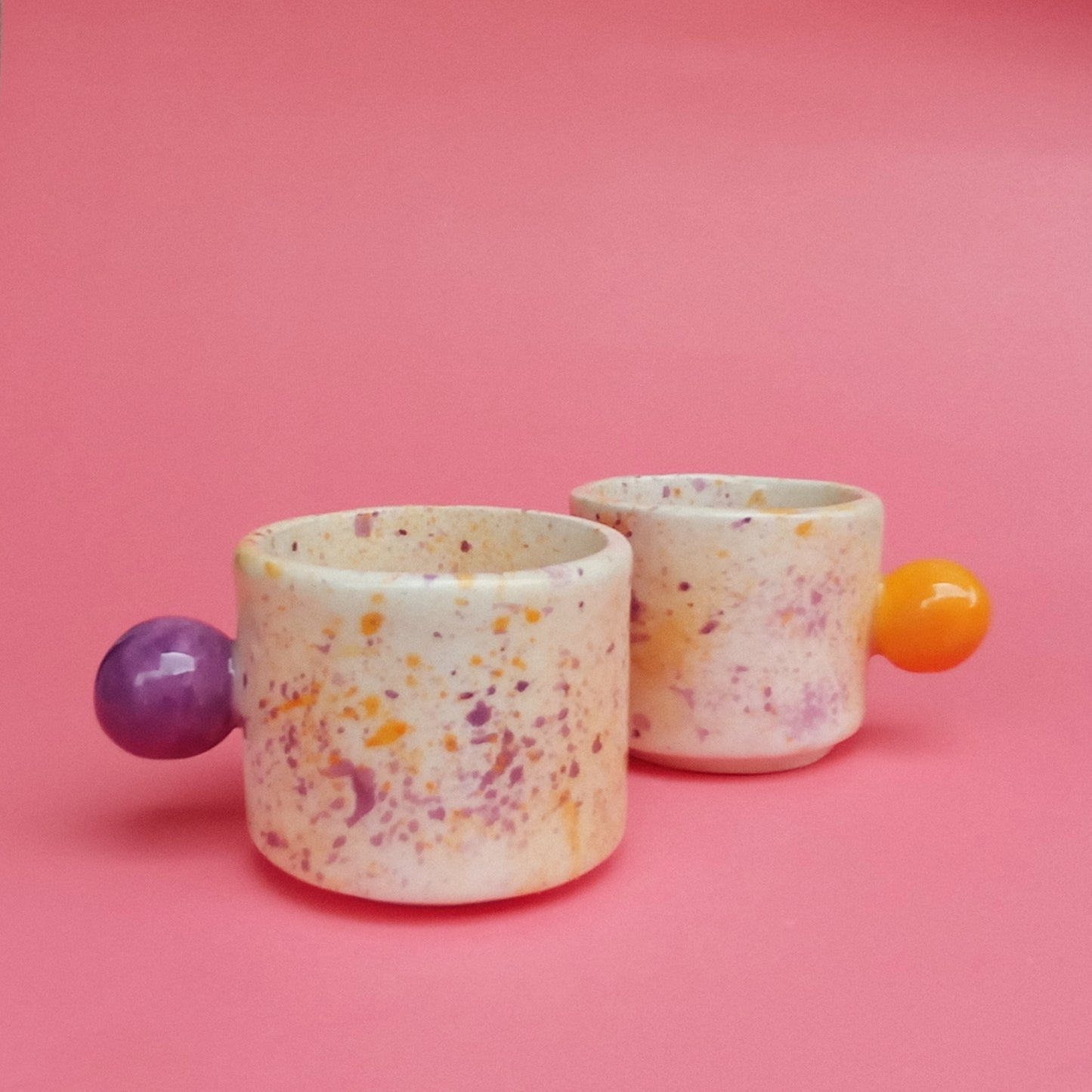Two ceramic cups with colorful speckles and spherical handles on a pink background