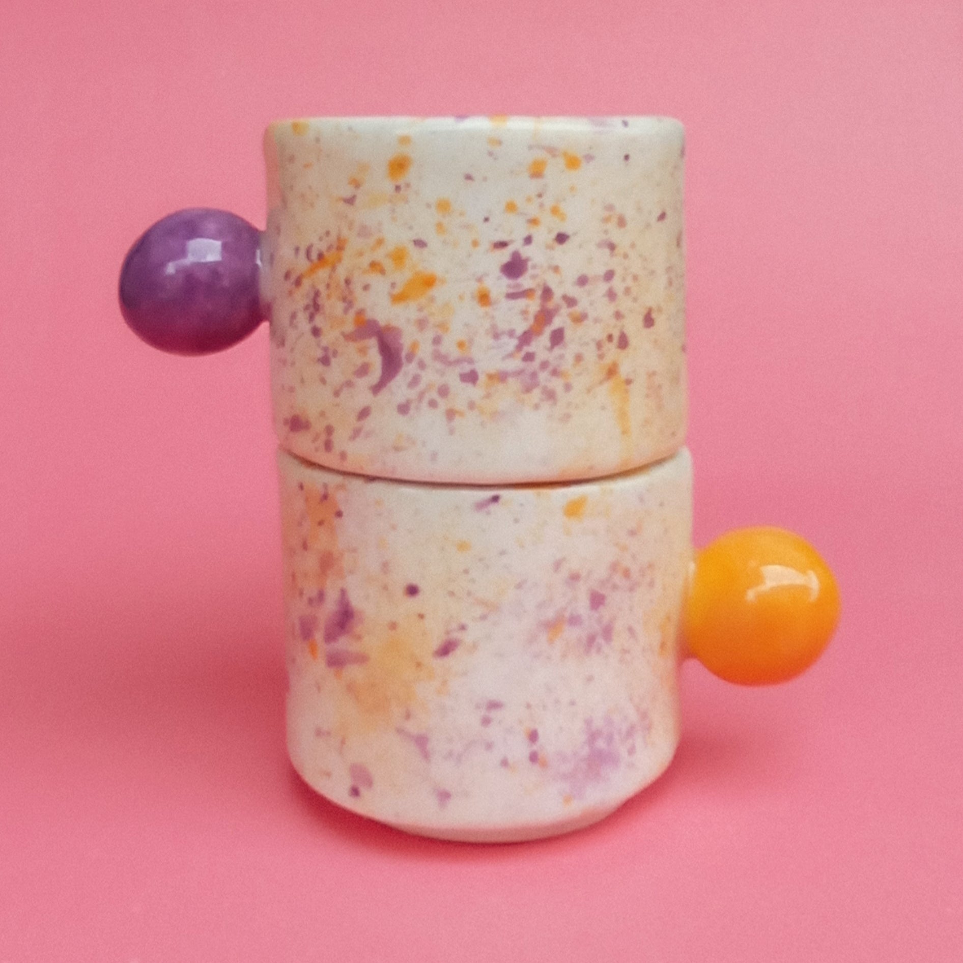 Speckled ceramic container with purple and orange handles on a pink background