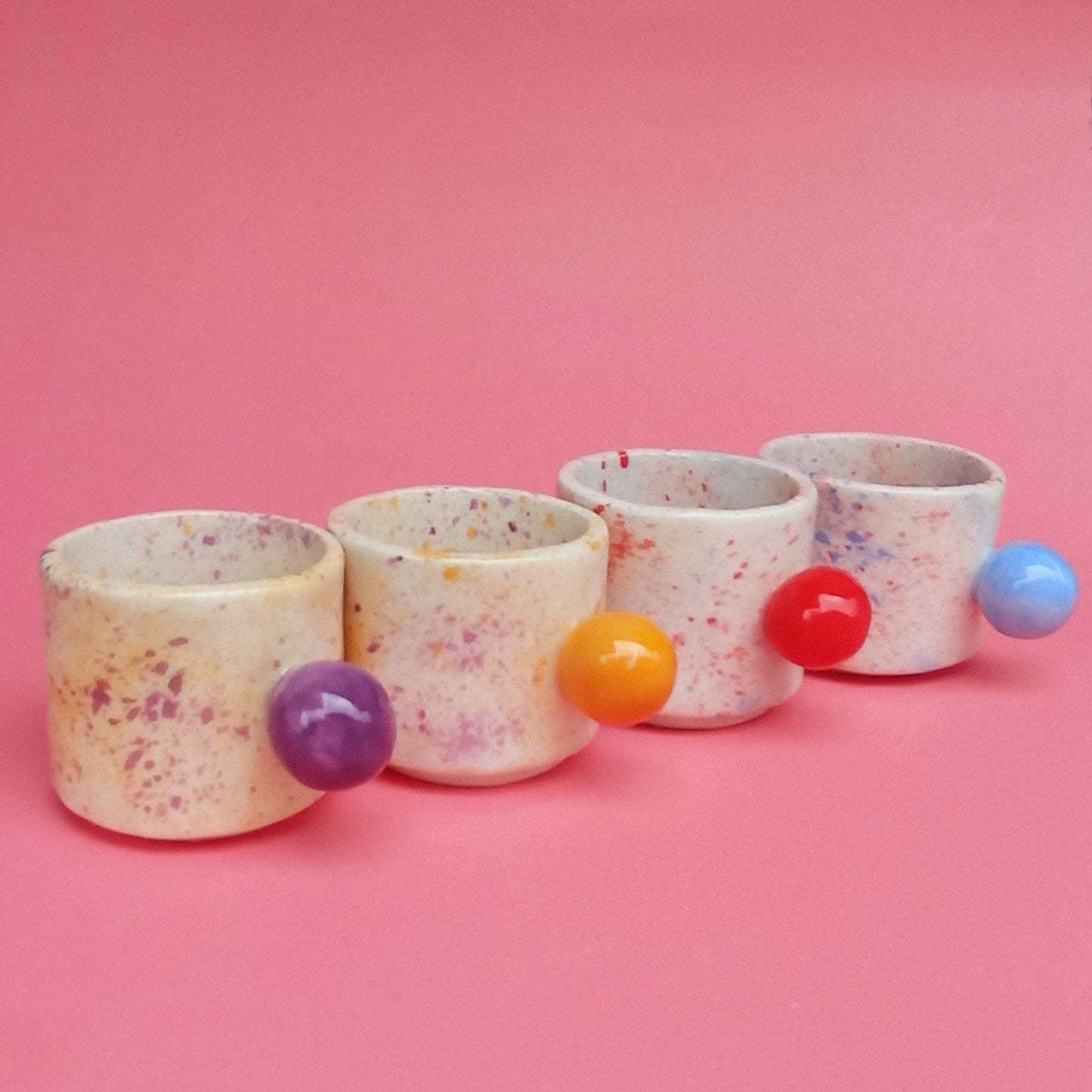 Four ceramic cups with colorful spherical handles on a pink background